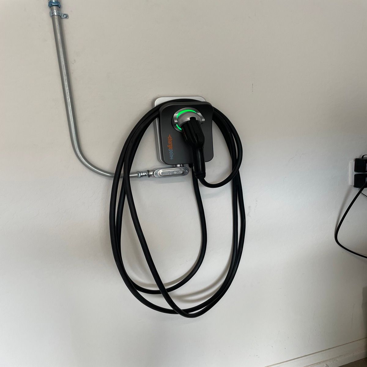 A ChargePoint electric vehicle charger is mounted on a white garage wall, connected through metal conduit, with its charging cable neatly wrapped and green indicator light showing operational status clearly.
