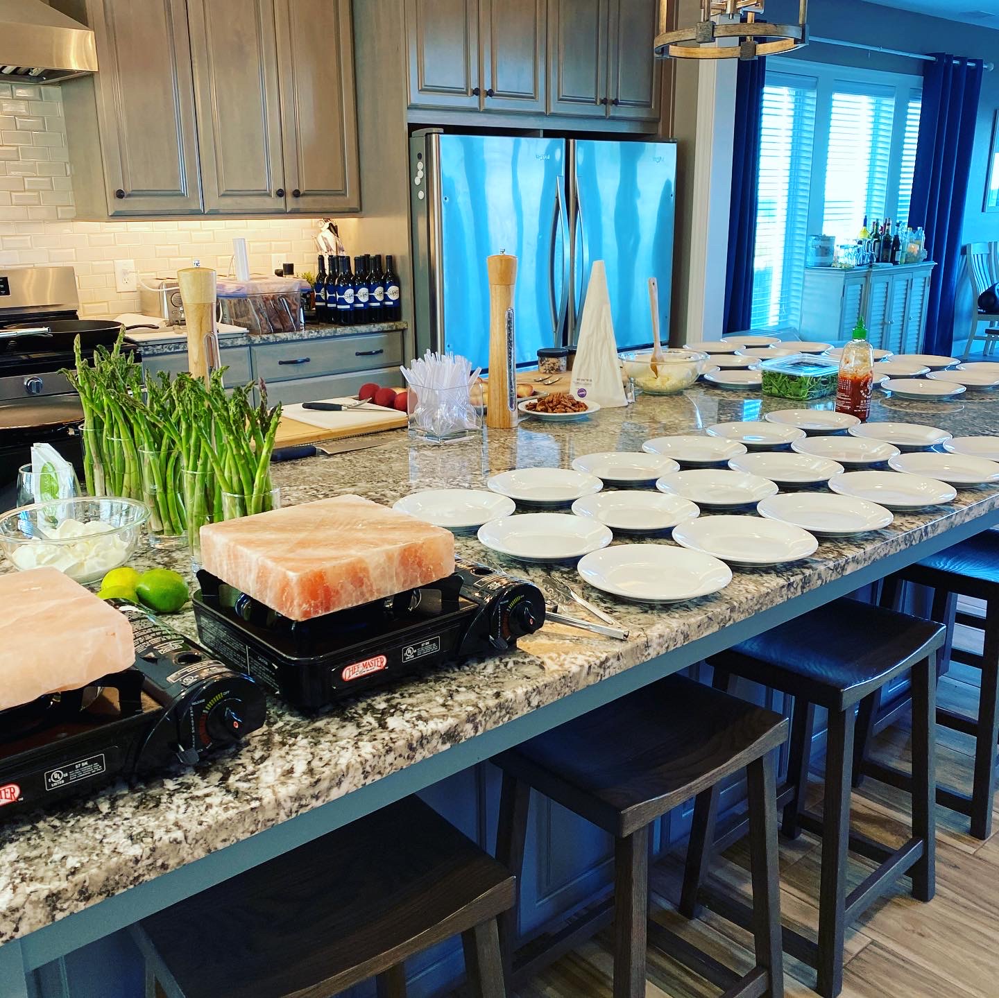 A kitchen counter is set up for a meal with many white plates stacked and spread out, a large block of pink salt on a portable stove, bunches of asparagus, bowls of food, and bottles of sriracha sauce. Several dark wooden bar stools are visible in the foreground, and the kitchen features grey cabinets and a stainless steel refrigerator. The scene appears to be a preparation for a gathering or a meal service.