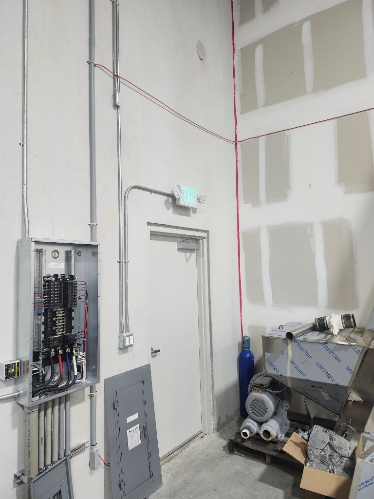 Industrial interior showing an open electrical breaker panel mounted on a wall beside a door, with exposed conduit wiring, exit sign lighting, and nearby mechanical equipment inside a utility workspace.