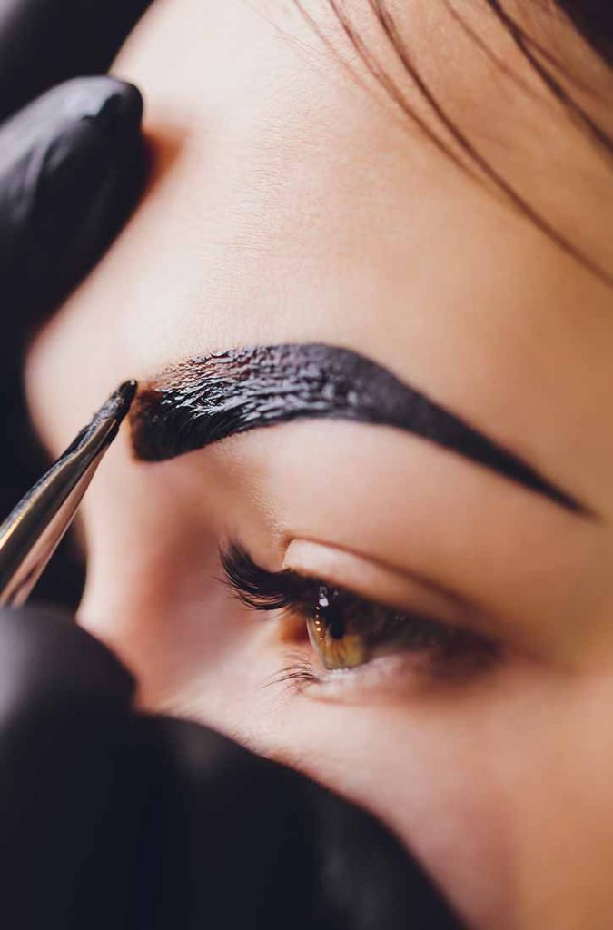 A close-up shot shows a person's eye and eyebrow being groomed with tweezers and a dark coloring product. The person is wearing black gloves, and the eyebrow is thick and appears to be undergoing a tinting or dyeing process.