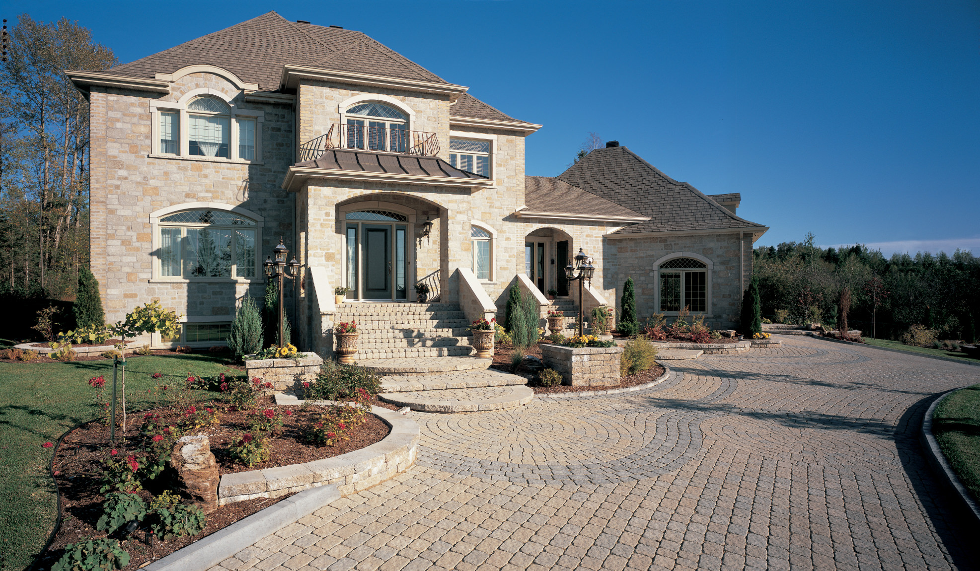 Large stone luxury home with arched windows, grand entry steps, landscaped flower beds, and a wide curved cobblestone driveway under blue skies.