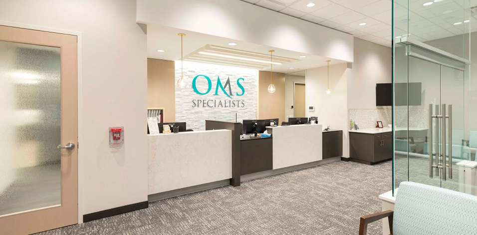 A spacious, modern healthcare clinic waiting area and hallway with wood-look laminate flooring, neutral gray walls, a reception desk, flush ceiling panel lights, and flat-screen monitors.