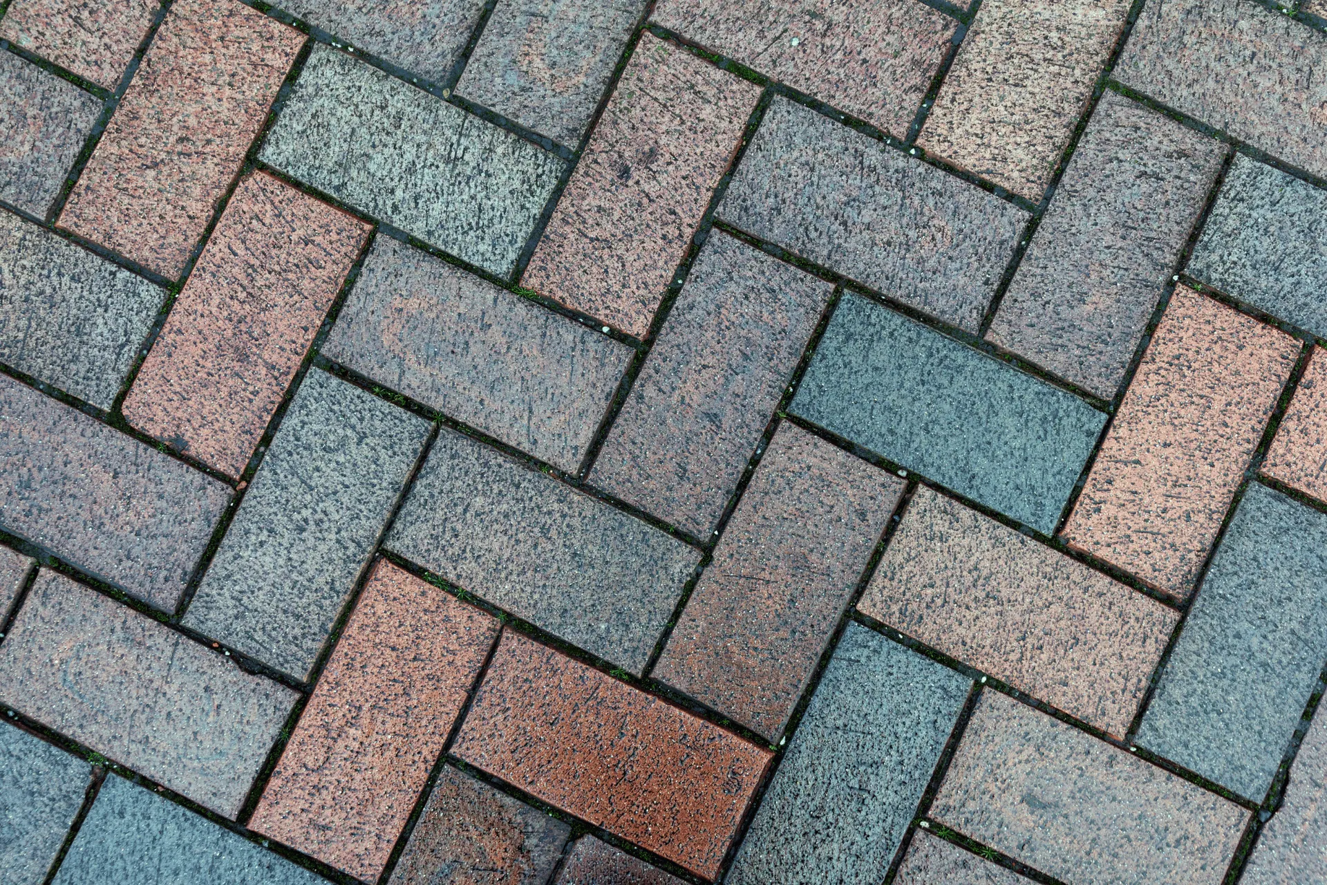 A close-up, overhead view of a herringbone pattern of brick pavers in muted red and gray tones. Small patches of green moss or grass are visible in the gaps between the bricks.