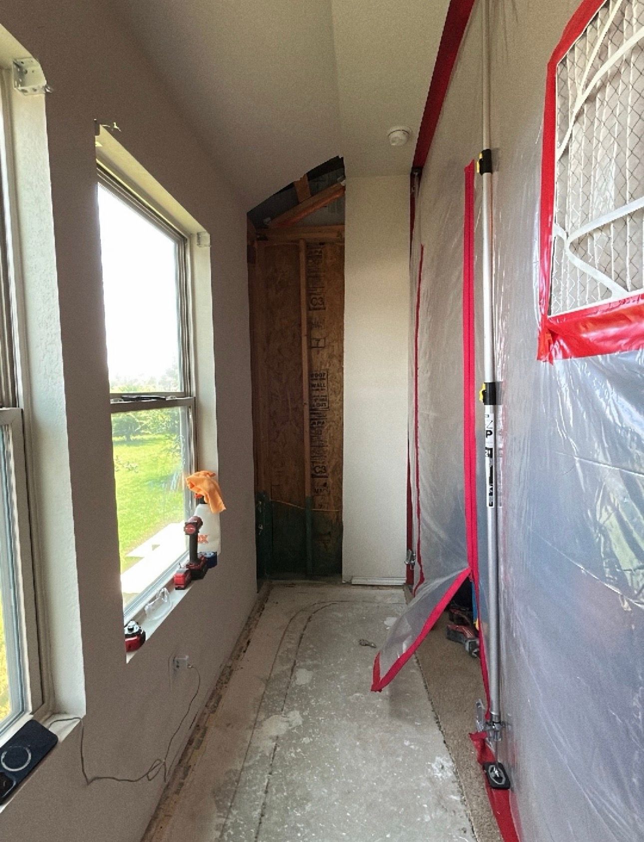A narrow residential hallway shows removed flooring and an opened wall section exposing framing, whi 
