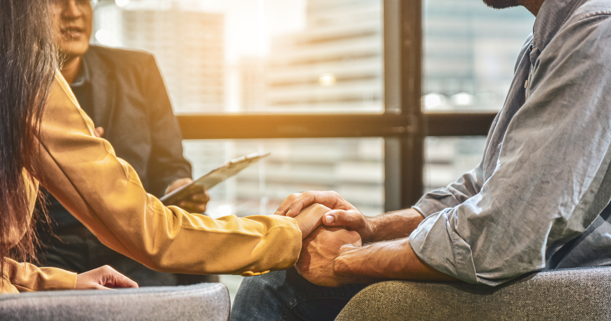 A close-up shot of three people holding hands while sitting in a meeting or therapy session. The person on the left is wearing a yellow jacket and reaching out to a person in the middle who is holding a clipboard. The person on the right is wearing a blue shirt and is being comforted by another person.