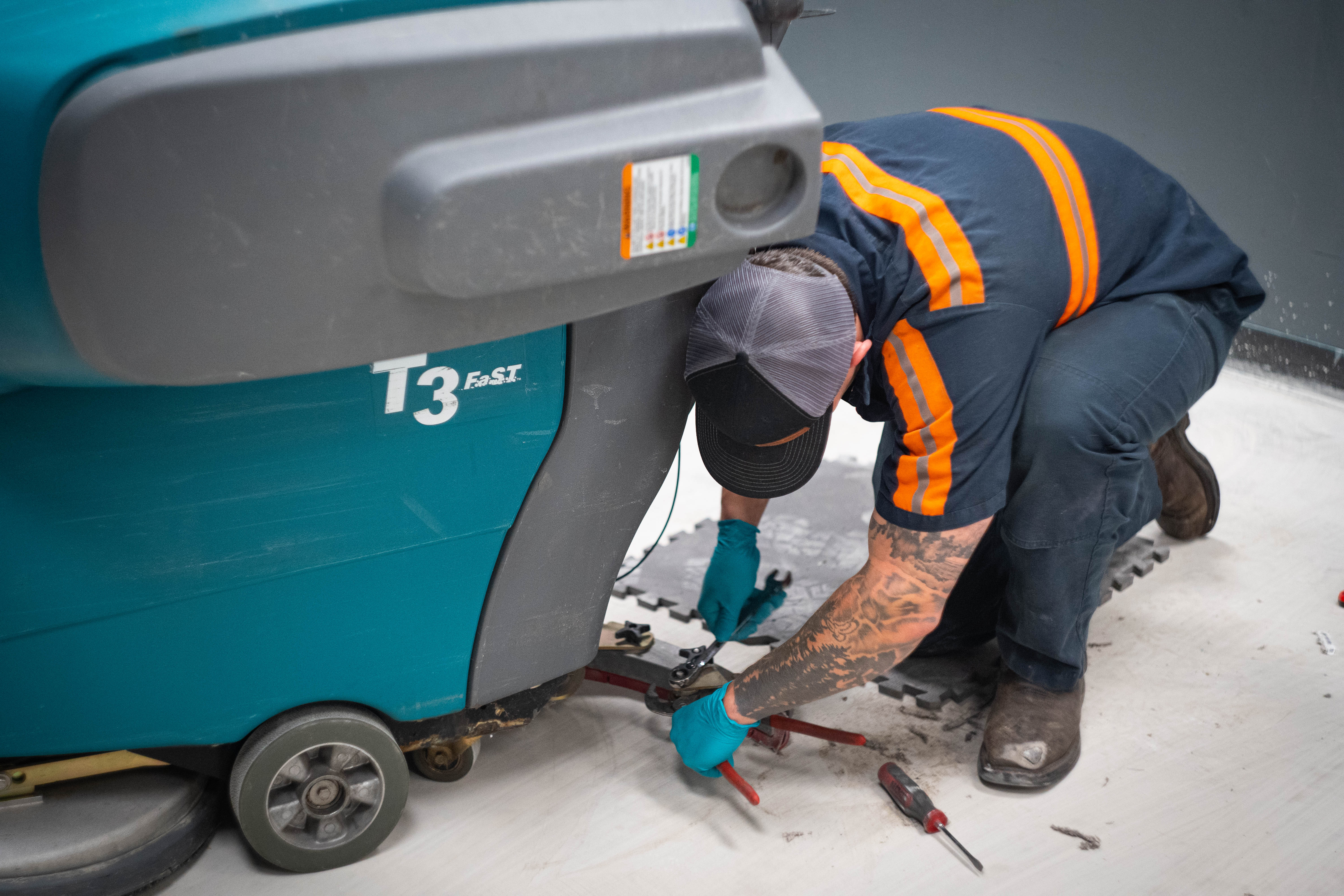 A WaySolutions field technician uses heavy-duty hand tools to perform lower mechanical repairs on a teal Tennant T3 FAST commercial floor scrubber. By performing precision maintenance on the undercarriage and squeegee linkage, our Layton-based technicians ensure supermarket and retail cleaning fleets operate at peak efficiency and leave floors perfectly dry.