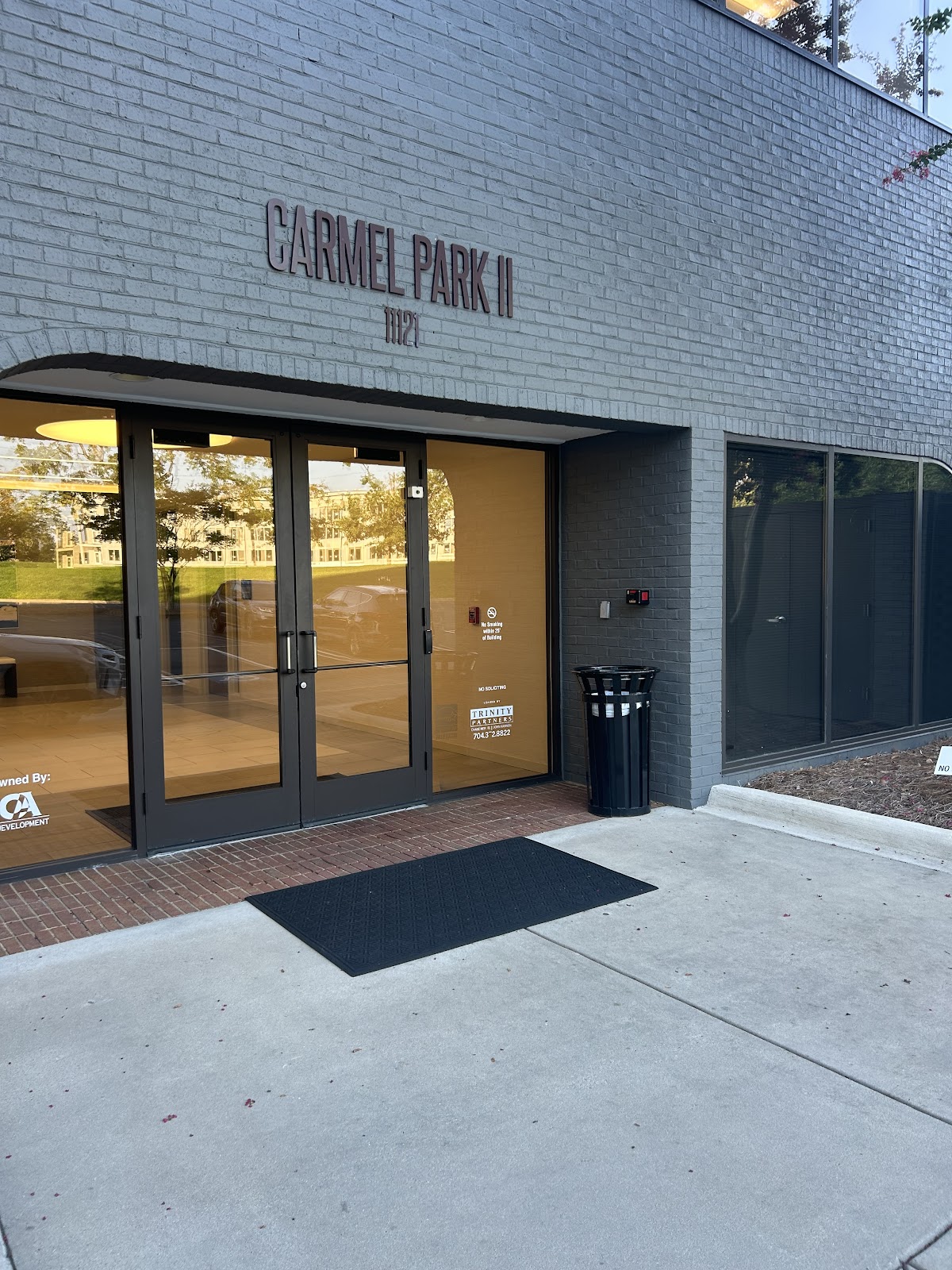 A close-up shot of the entrance to a building called CARMEL PARK II, with the address 1121 above the double glass doors. The building is made of dark gray brick. The doors lead into a well-lit interior. A black trash can stands to the right of the entrance, and a black doormat is placed in front of the doors.