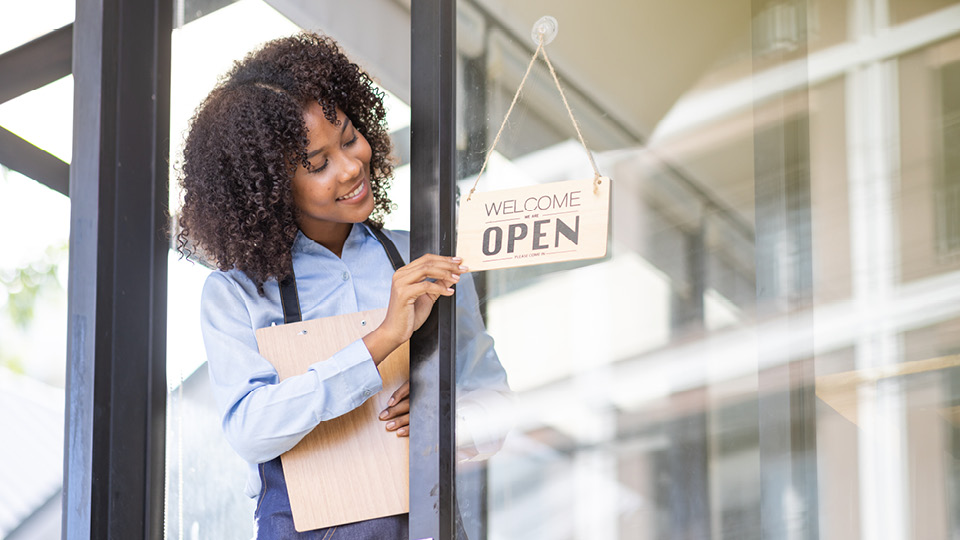 Woman holding a clipboard opens a glass door with a hanging sign that reads 'Welcome Open'.