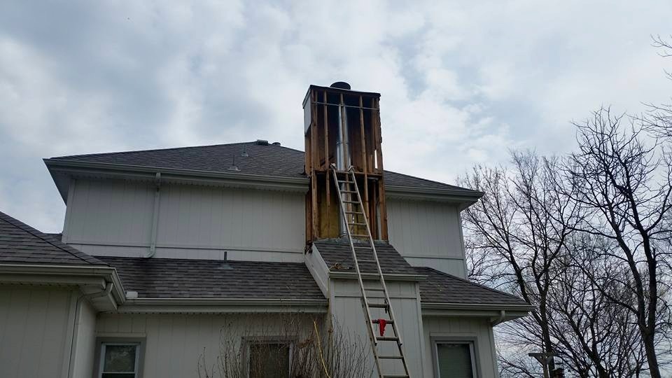 A residential home exterior is undergoing wood rot repair on the chimney structure. The photo shows damaged and exposed wood framing being repaired with a ladder set up for access, demonstrating professional restoration work in progress. Ideal for showcasing wood rot repair services, structural restoration, and exterior home maintenance.