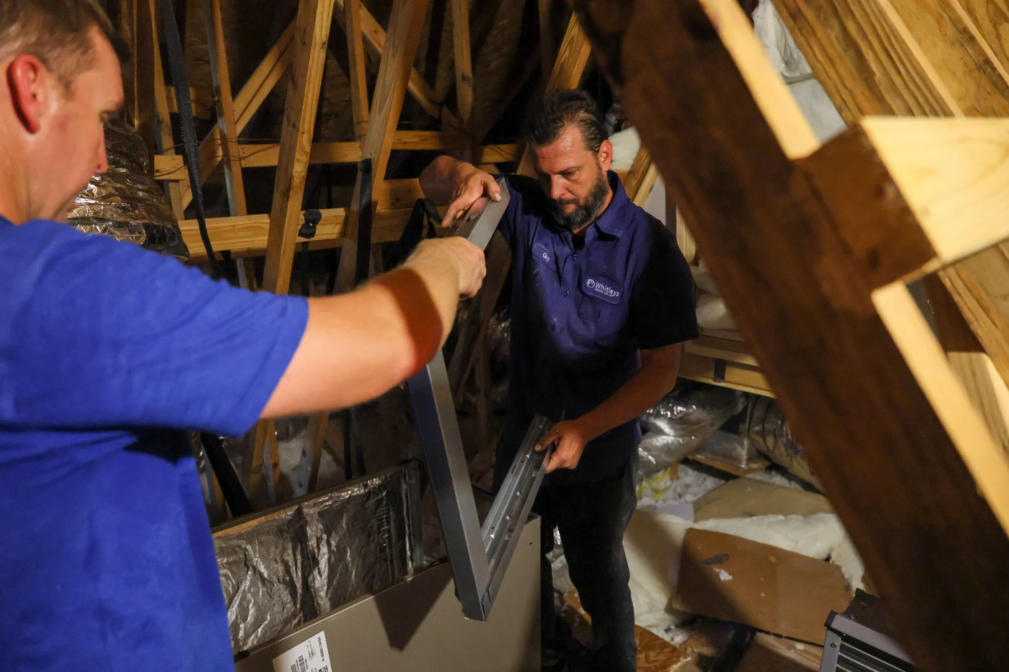 Two men are working in an attic, assembling a metal frame. The man on the left is wearing a blue t-s 