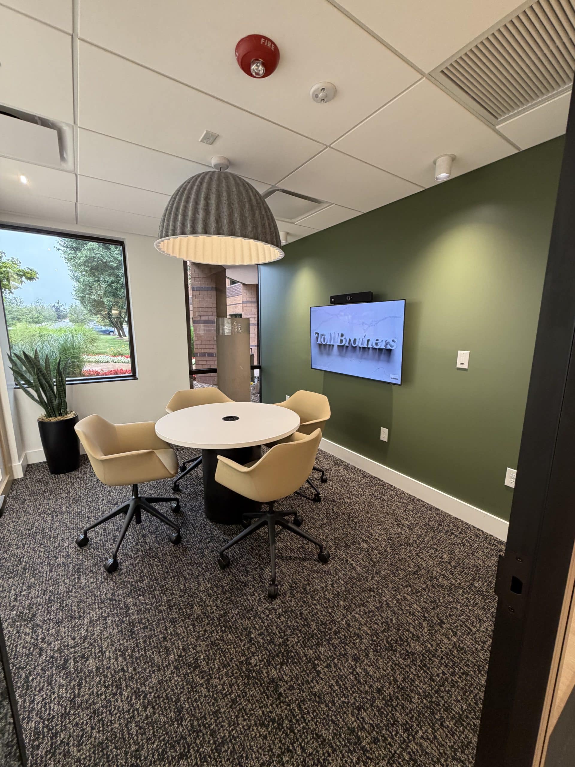This meeting room at the Toll Brothers Michigan Division Office & Design Studio brings comfort, technology, and distinctive finishes together within Schonsheck, Inc.’s commercial construction portfolio. An olive-green accent wall, upholstered seating, and a statement pendant light create an inviting setting for small group discussions. A wall-mounted display and conferencing equipment complement the room’s practical layout, while natural light adds warmth to the professional office interior.