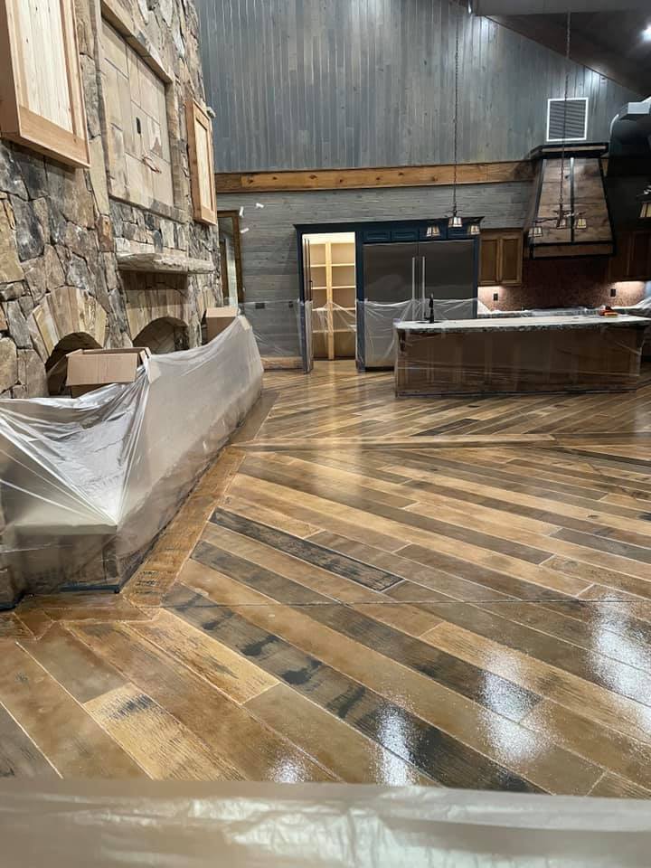 A spacious room with a large stone fireplace on the left, and a kitchen area with stainless steel appliances and a large island on the right. The floor is made of wooden planks laid in a chevron pattern and appears to be newly finished or polished, with protective plastic sheeting visible in the foreground. The walls are a mix of stone and wood paneling, and the ceiling is high with exposed wooden beams and industrial-style light fixtures.