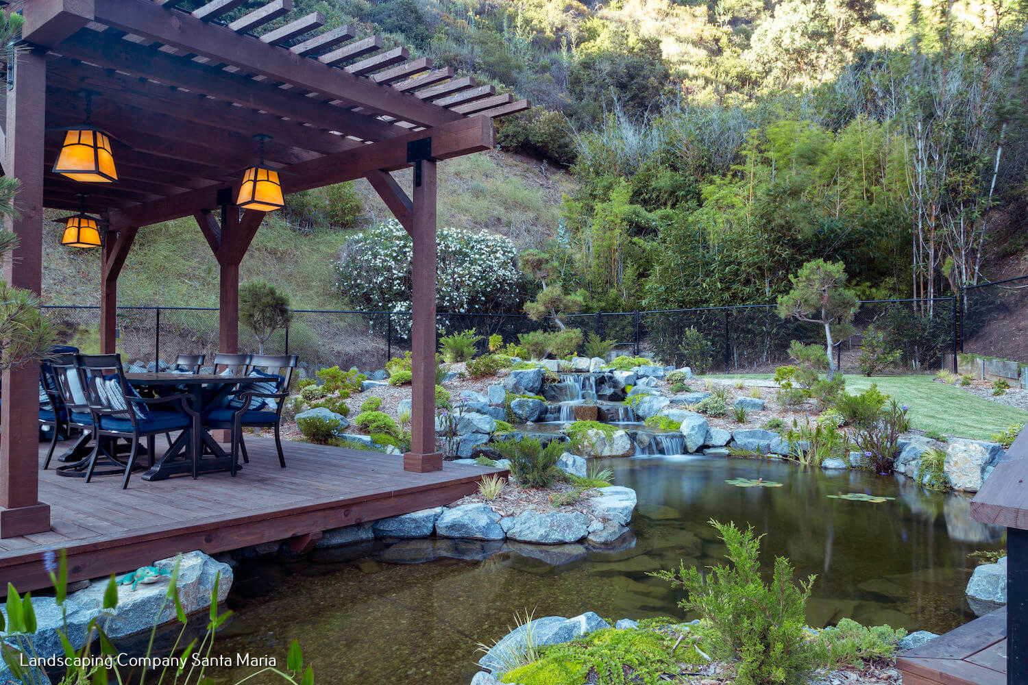 Wooden pergola with glowing lanterns and outdoor dining set overlooks a tranquil koi pond, rocks, waterfall, and lush garden hillside.
