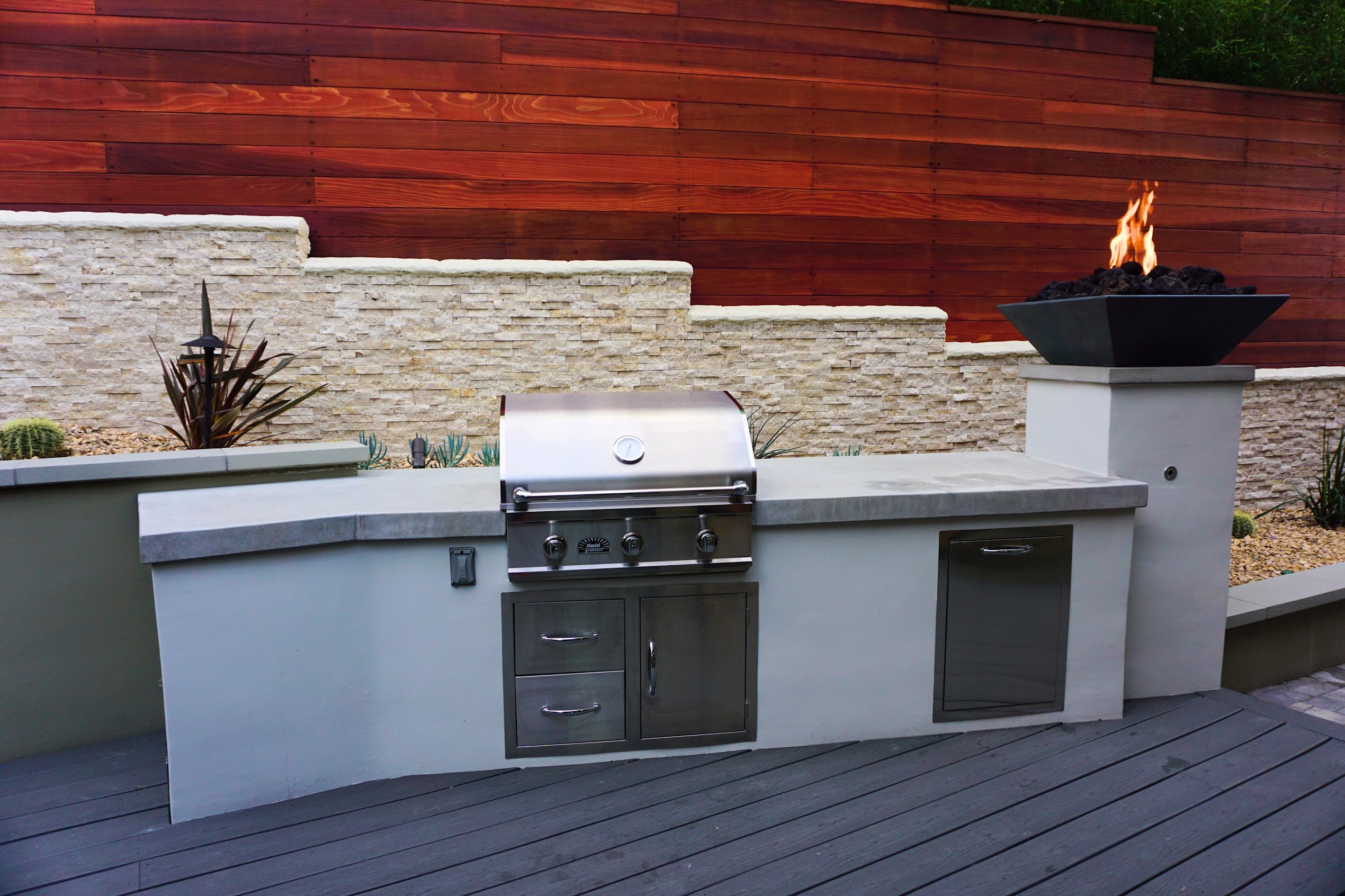 Modern outdoor kitchen with stainless-steel grill, gray counters, stone wall, red wood fence, plants, and a flaming fire bowl.