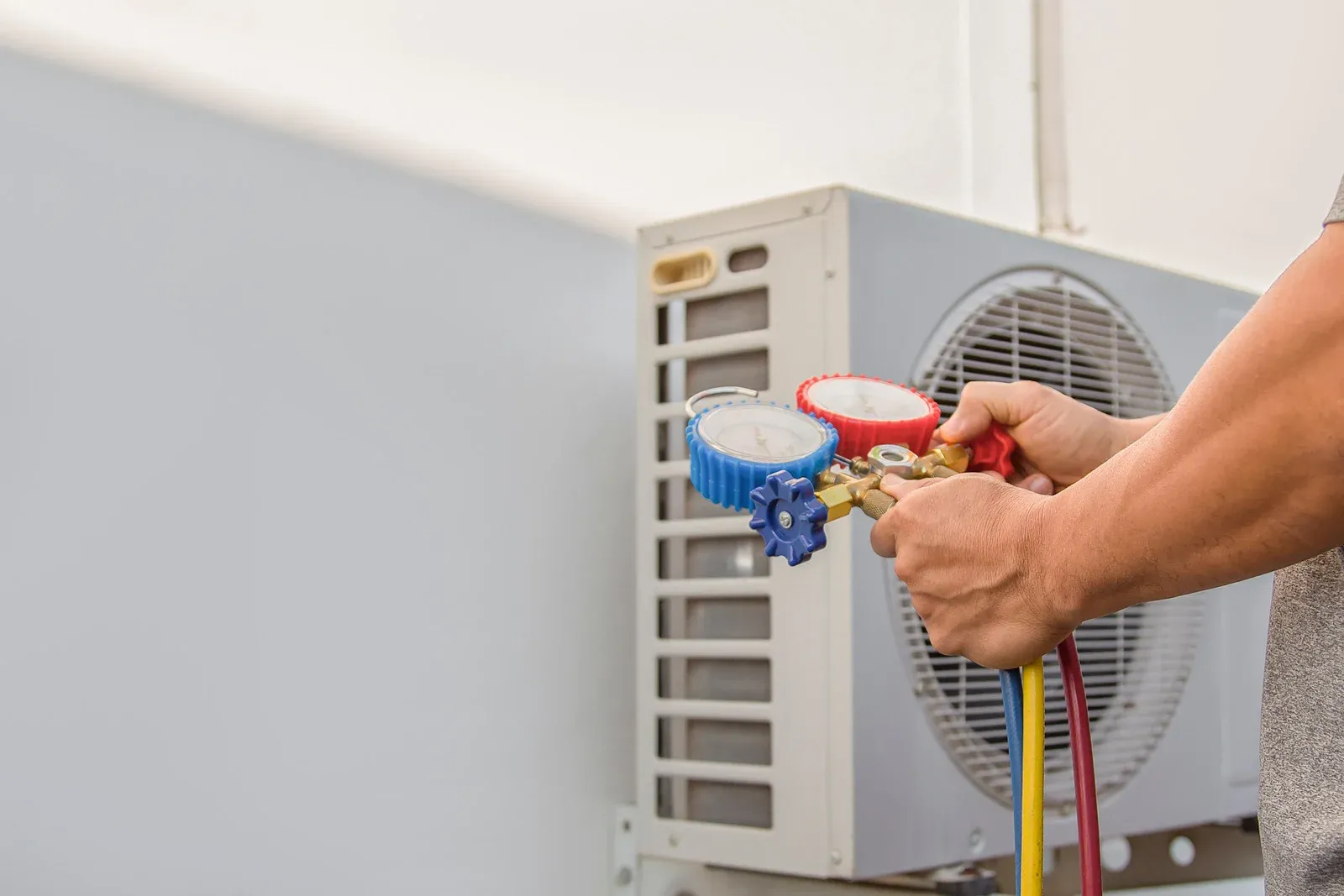 A technician holds HVAC manifold gauges with red, blue, and yellow hoses while inspecting an outdoor air conditioning unit, performing maintenance and checking refrigerant pressure for proper cooling performance.