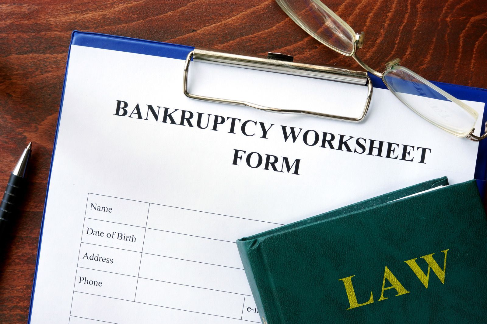 A bankruptcy worksheet form with fields for name, date of birth, address, and phone rests on a blue clipboard, with a green law book, eyeglasses, and a black pen nearby.