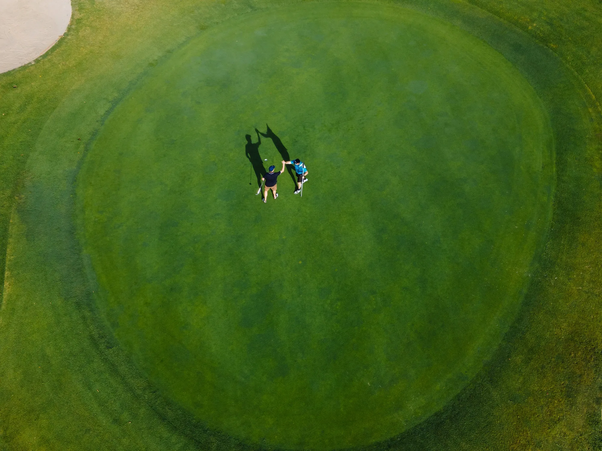 An aerial, high-angle shot of two golfers celebrating with a high-five on a vibrant green golf course. One golfer is wearing a blue shirt and shorts, the other is wearing a black shirt and shorts with a blue cap. They are standing on the putting green, with their shadows cast long behind them. To the left of the frame is a sand trap, and the varying shades of green on the course indicate the different mowing patterns and contours of the terrain. The image captures a moment of sportsmanship and enjoyment on a sunny day.