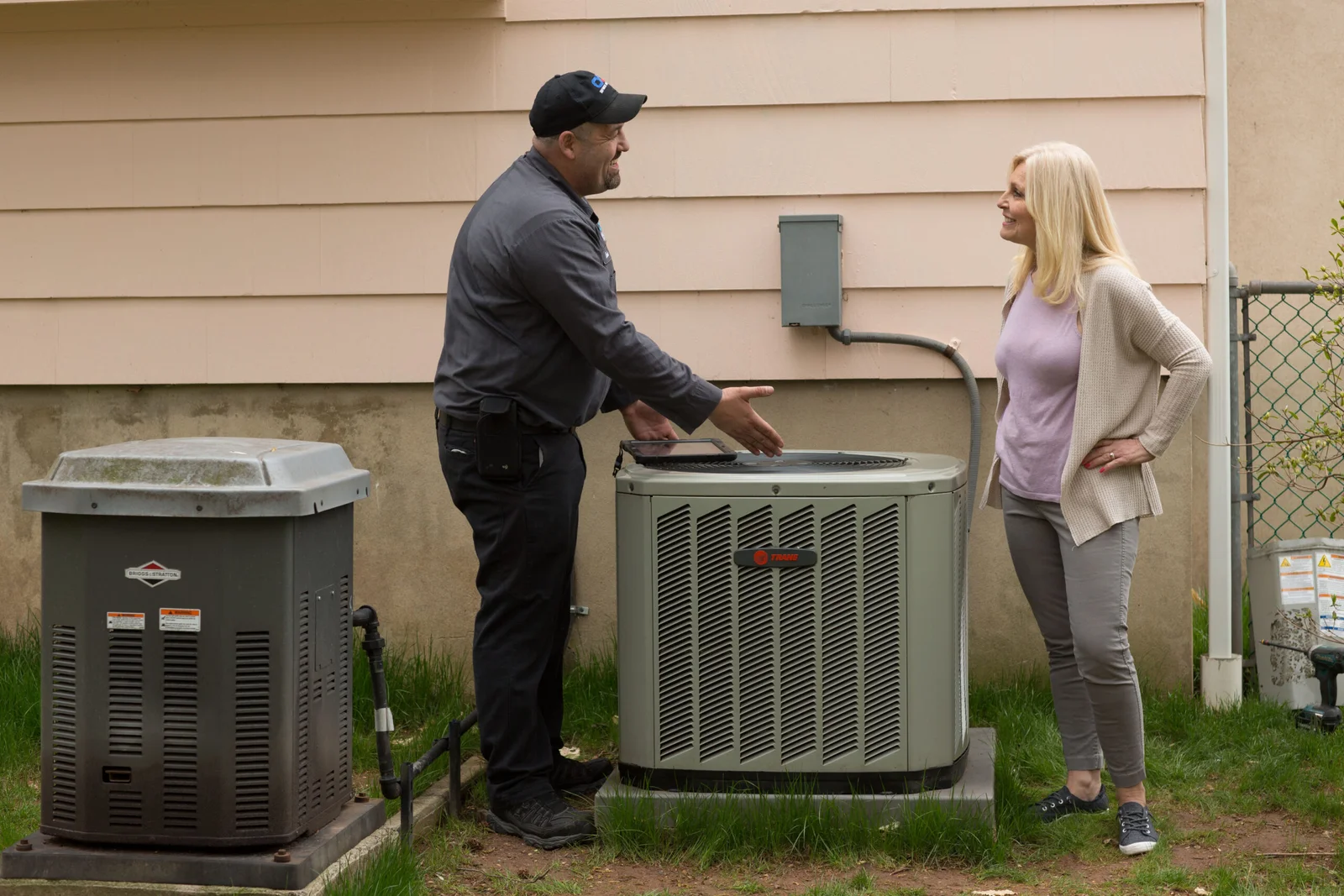 An HVAC technician in a gray uniform and a blue cap is showing a tablet to a woman in a tan cardigan and a lavender shirt, standing in front of two large air conditioning units on the side of a house.