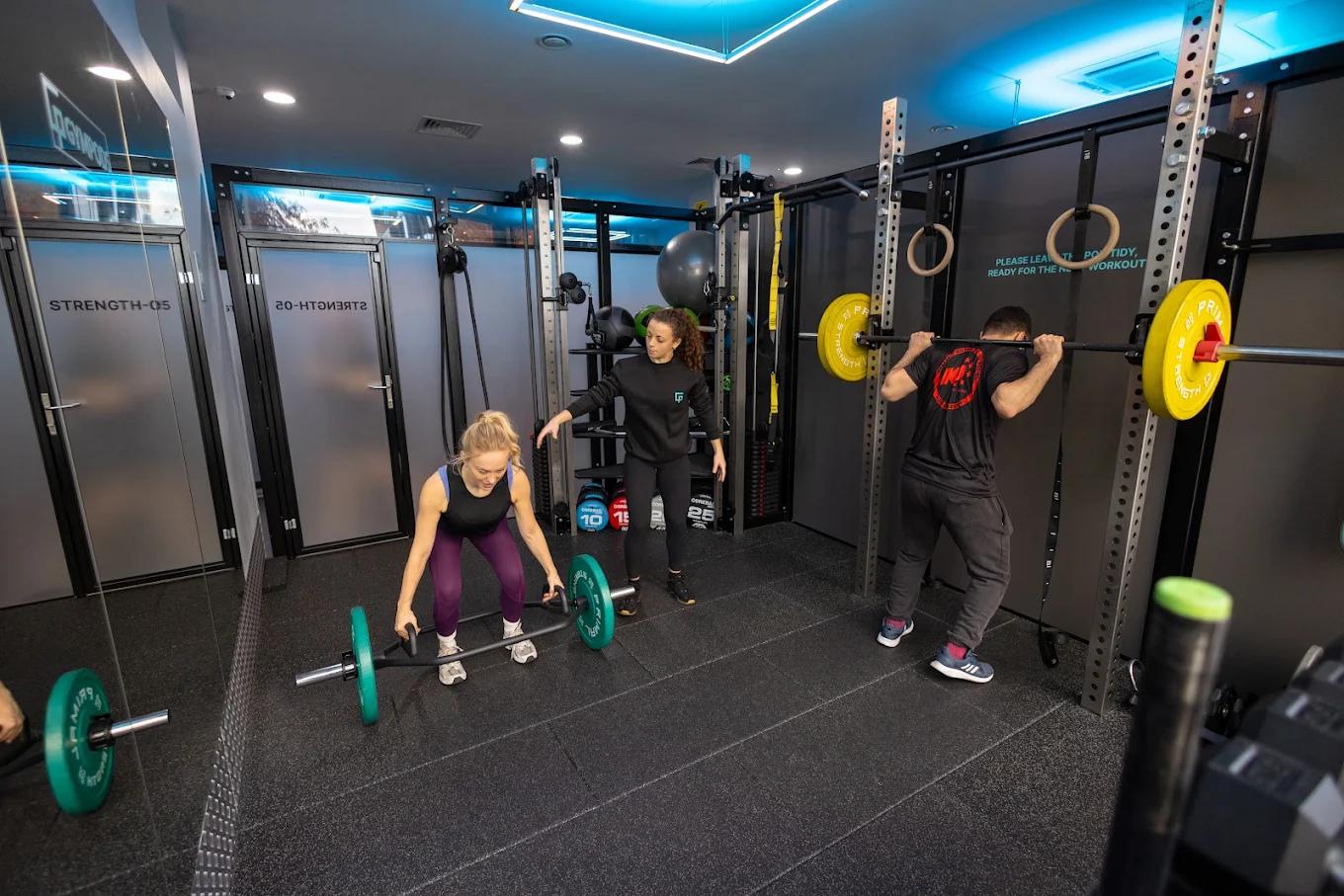 Personal trainer coaching two people lifting barbells in a private Strength POD at GYMPODS Putney.