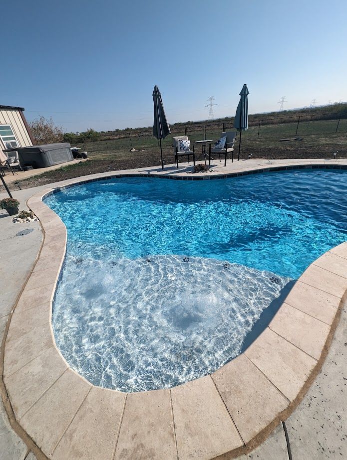 A finished freeform swimming pool features sparkling blue water and a large tanning ledge with bubbling bubblers. The surrounding stone coping and concrete deck overlook a wide, open landscape under a clear sky.