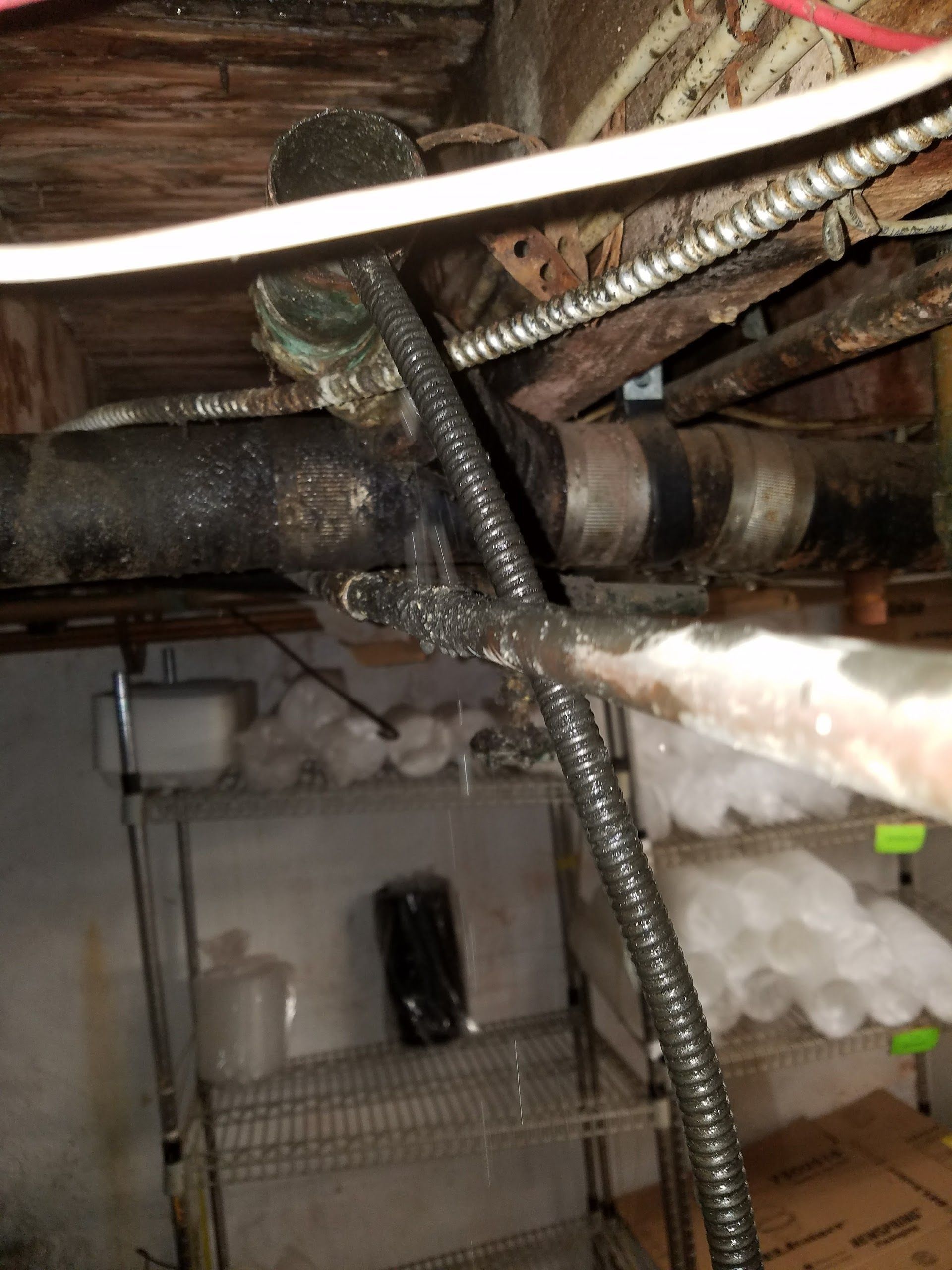 Exposed basement plumbing with corroded pipes and visible leak dripping water, showing deteriorated connections and buildup, indicating urgent need for professional repair, maintenance, and leak detection to prevent further damage.