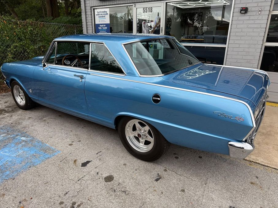 A beautiful, light blue classic Chevrolet Nova with chrome accents and aftermarket alloy wheels is parked outside an automotive service center. The car is professionally detailed and in excellent condition.