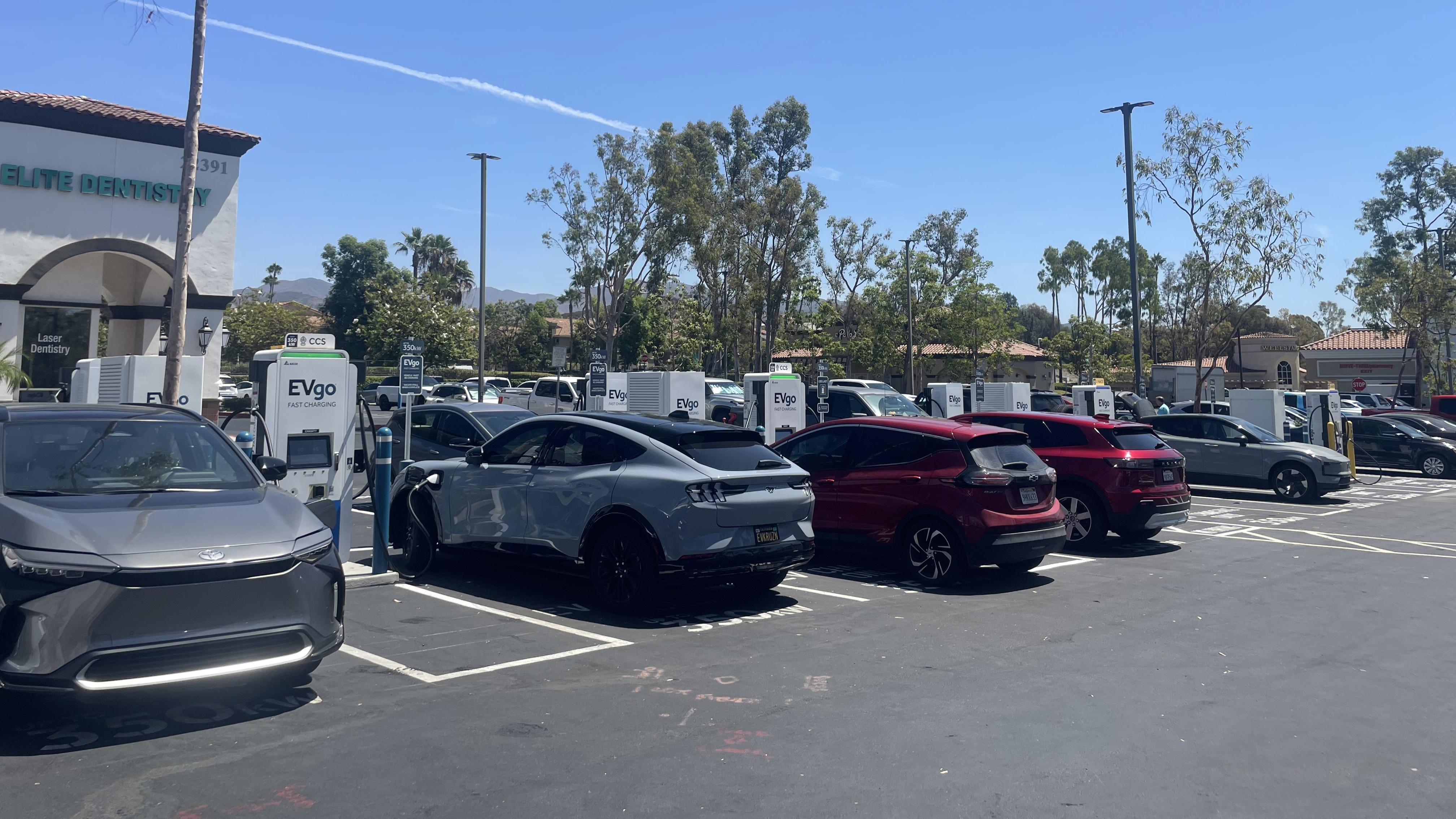 Several EVgo fast charging units installed in marked parking spaces at a public charging station, with EVgo logos on the chargers, protective bollards around each unit. NACS and CCS chargers featured in the image.