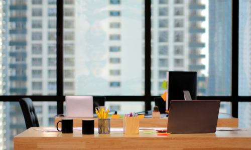 Modern office desk setup with laptops, mugs, and stationery, overlooking a city skyline through large windows.