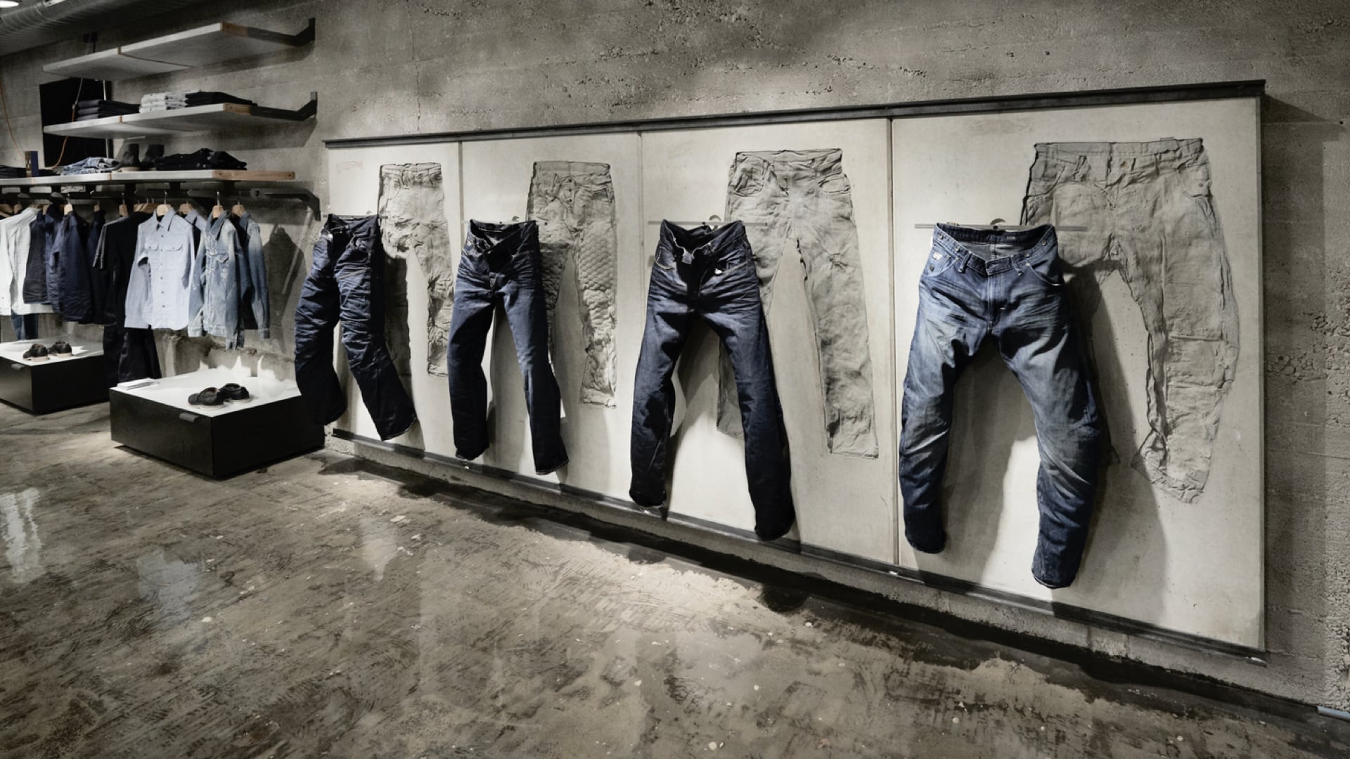 Des jeans sont suspendus contre un mur de ciment industriel, à côté de chemises bleues boutonnées.