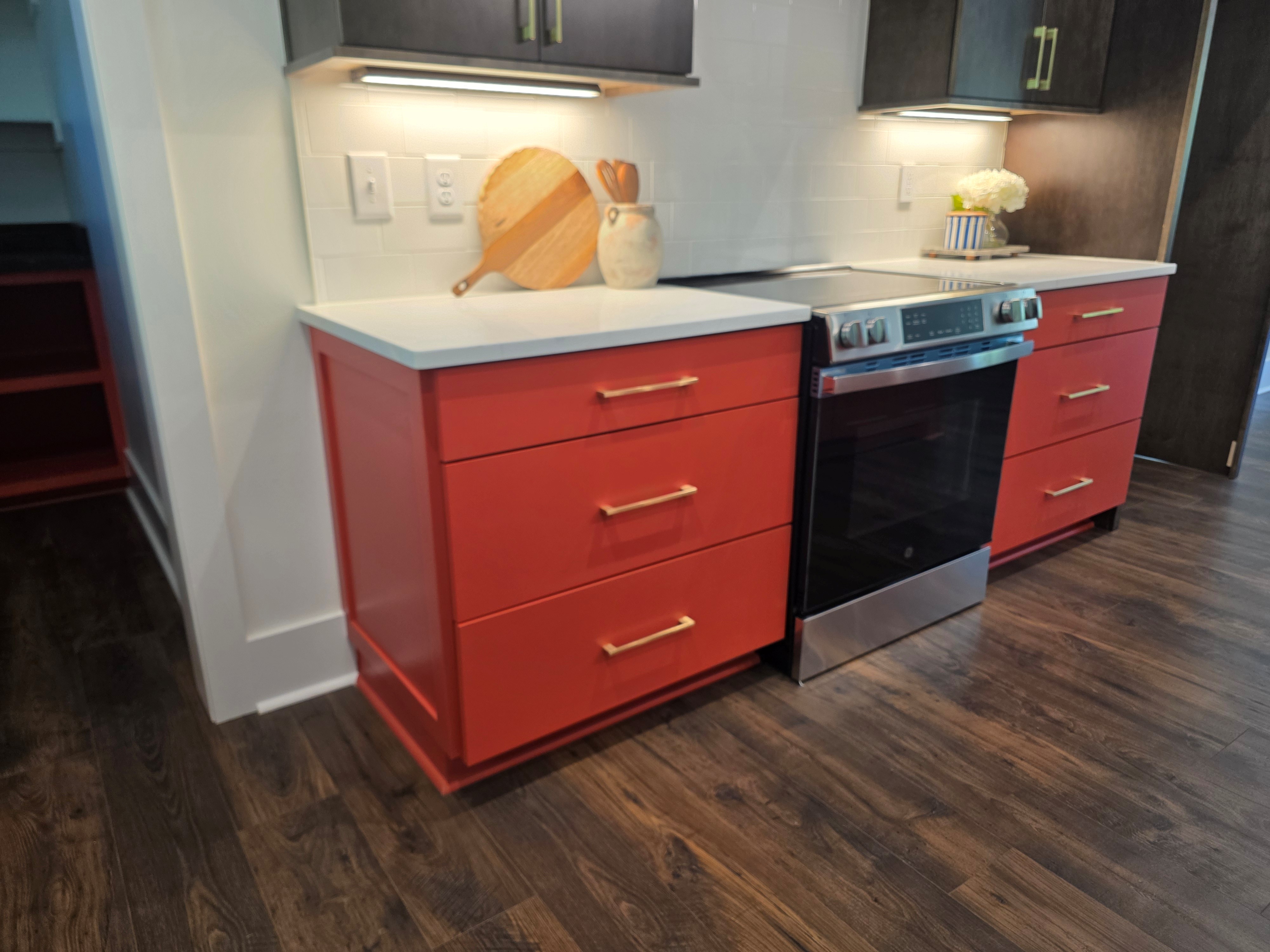 Modern kitchen with bold red lower cabinets featuring gold hardware, white quartz countertops, dark upper cabinets, and a stainless steel oven with under-cabinet lighting.