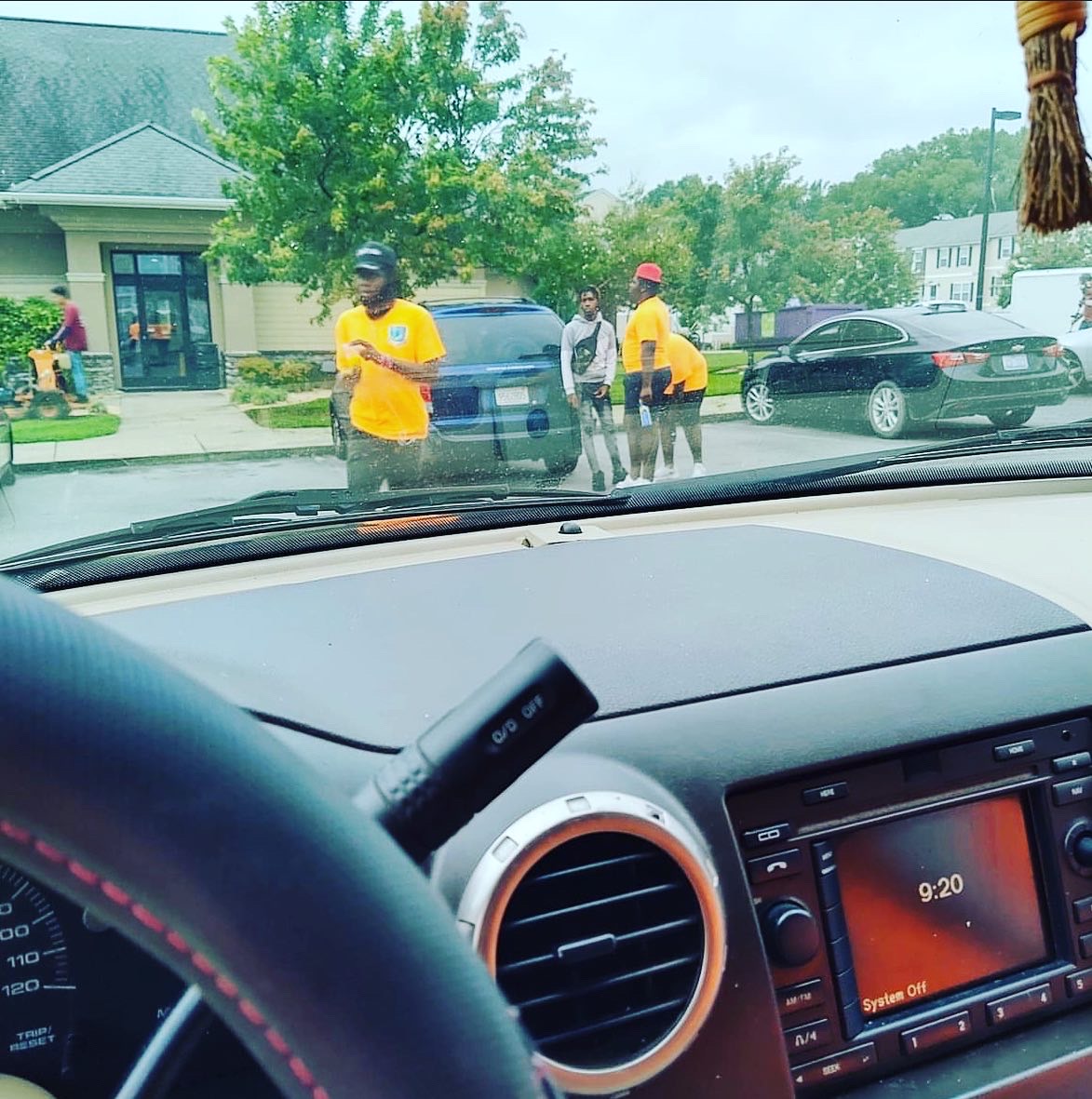 A group of young people wearing bright yellow shirts stand near cars in what appears to be a parking lot. One person is using a landscaping tool, possibly a leaf blower, on the left side of the frame. The perspective is from inside a car, showing the dashboard, steering wheel, and radio display which reads '9:20 System Off'. The weather appears overcast.