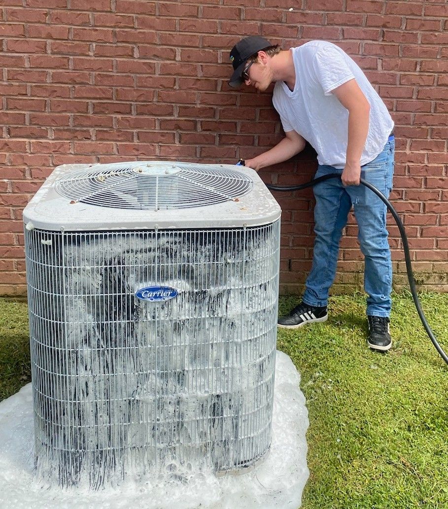 Technician cleaning outdoor air conditioning condenser unit with water hose, removing dirt and debris buildup, improving system efficiency, airflow, and performance, while maintaining residential HVAC equipment near brick exterior wall.