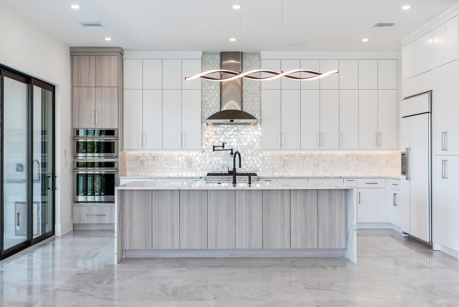 Bright custom kitchen with a large waterfall island, sleek white cabinetry, pendant lighting, stainless appliances, and an open modern layout.