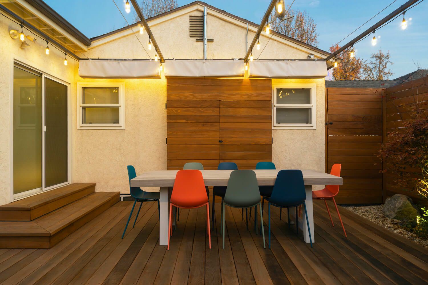 Warmly lit outdoor dining area on a wooden deck, with a long table and colorful chairs beside a modern home, creating a cozy evening atmosphere.