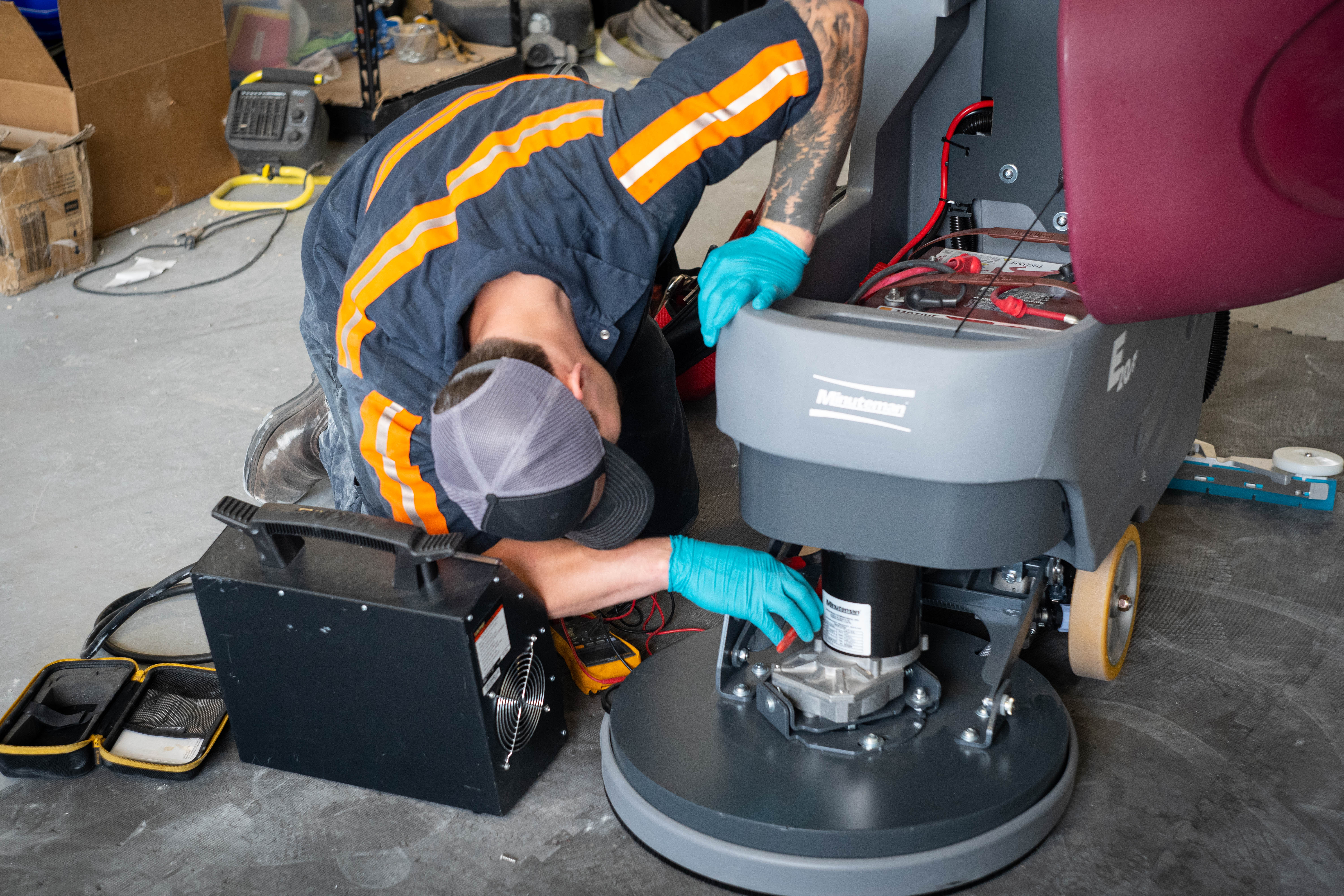 A WaySolutions service expert reaches deep into the lower brush deck of a Minuteman E20 automatic floor scrubber to perform critical mechanical diagnostics on the scrub motor. Surrounded by professional electrical testing equipment, the technician evaluates the drive system to guarantee maximum scrubbing pressure and efficiency for demanding commercial and industrial cleaning applications.