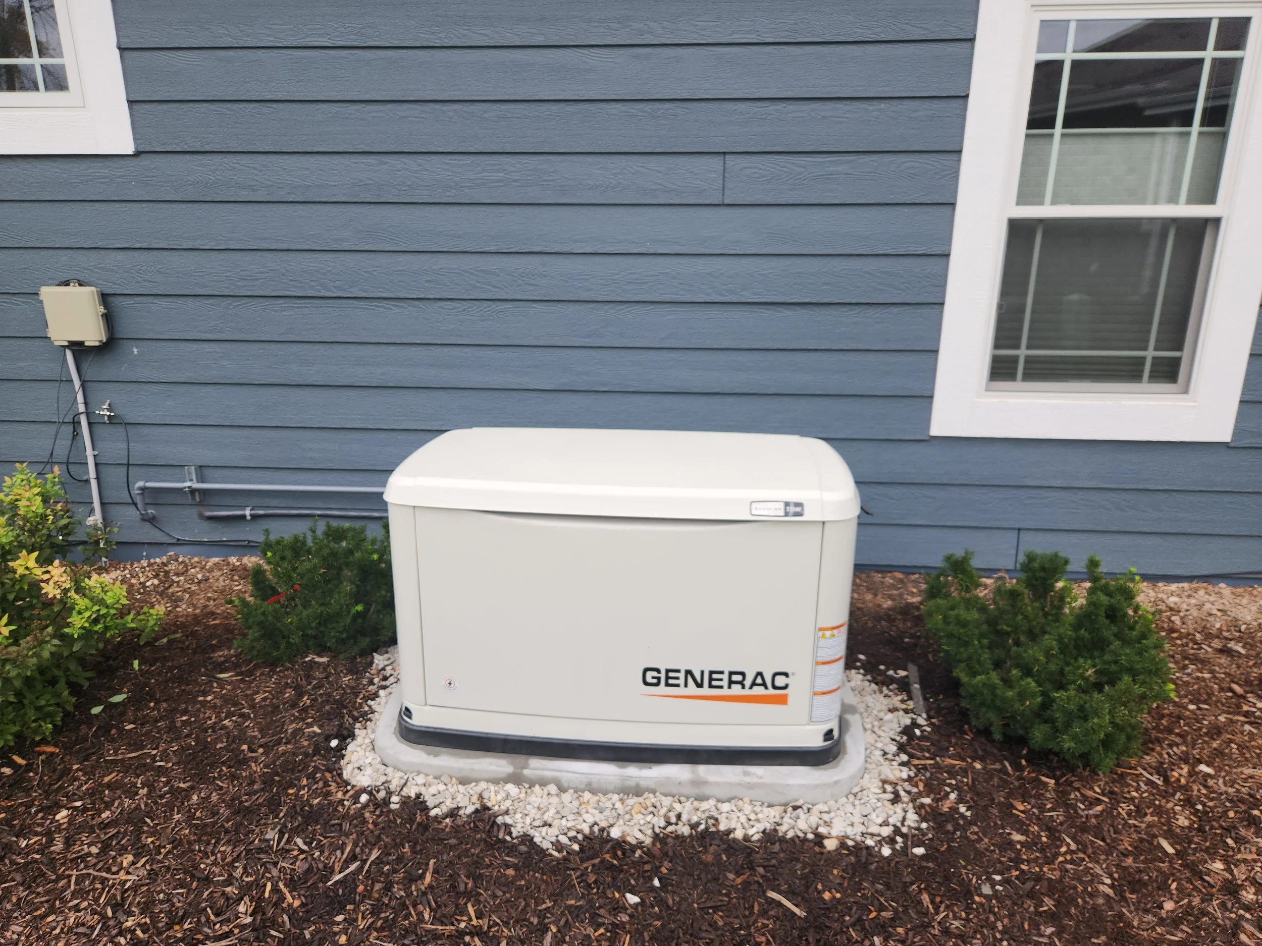A Generac brand generator is installed next to a blue siding house with a white-trimmed window. The generator sits on a concrete pad surrounded by mulch and small white rocks. There are also small green bushes around the generator and house.