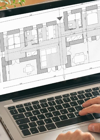A detailed, overhead shot focuses on a laptop screen displaying a black and white technical architectural floor plan of an apartment. A person's hands, with clear focus on the right hand, are shown interacting with the laptop's keyboard in the foreground, suggesting they are working on the design or plan.