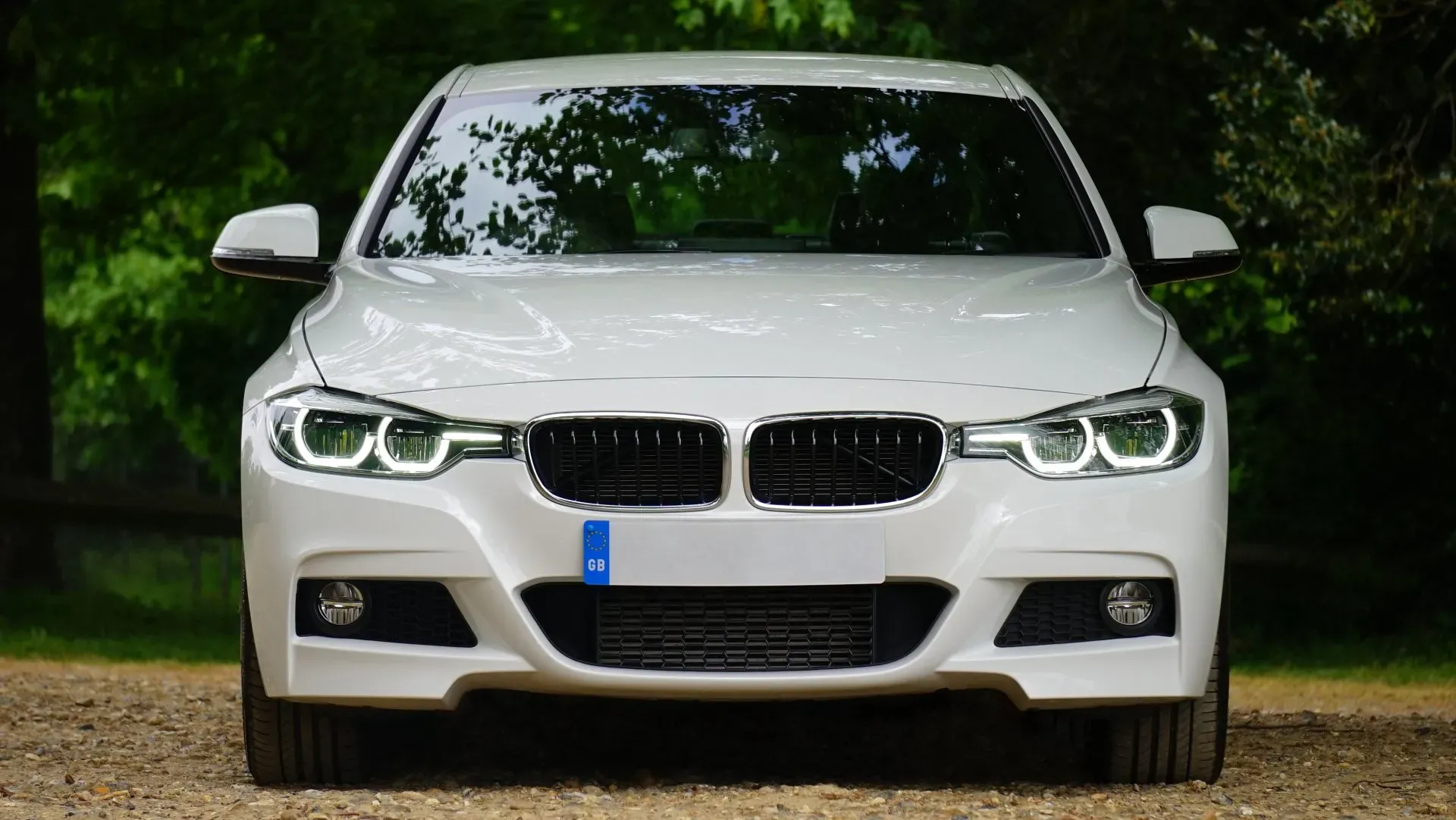 A white BMW sedan is parked on a gravel road with a dense forest in the background. The car's headlights are on, illuminating the front grille and bumper. The image is taken from a low-angle, front-facing perspective, emphasizing the car's powerful stance.