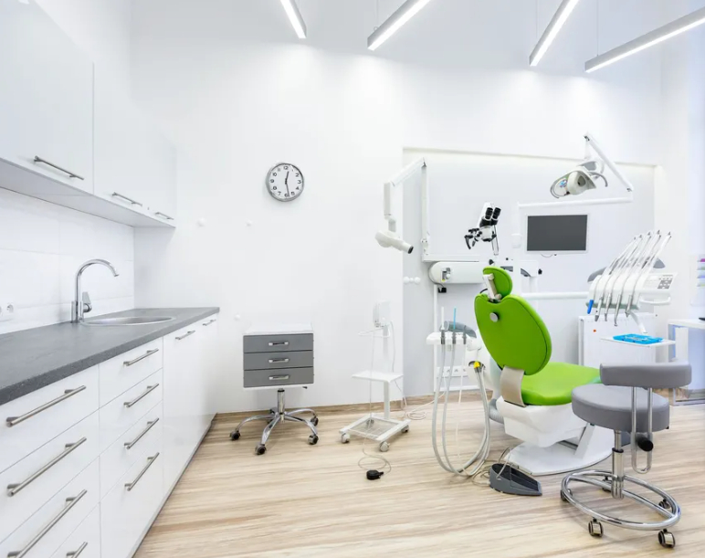 A brightly lit dental treatment room featuring a beige patient chair, overhead dental lamp, modern cabinetry, large floor-to-ceiling windows, and a wall-mounted digital x-ray display.