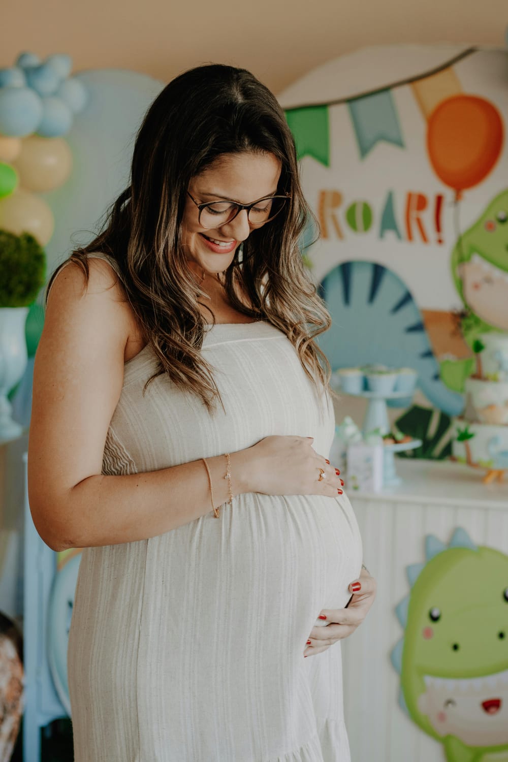 A pregnant woman smiles as she cradles her belly at a baby shower. She is wearing a light-colored, strapless dress and glasses. The background is decorated with a dinosaur theme, featuring balloons and banners that say "ROAR!".