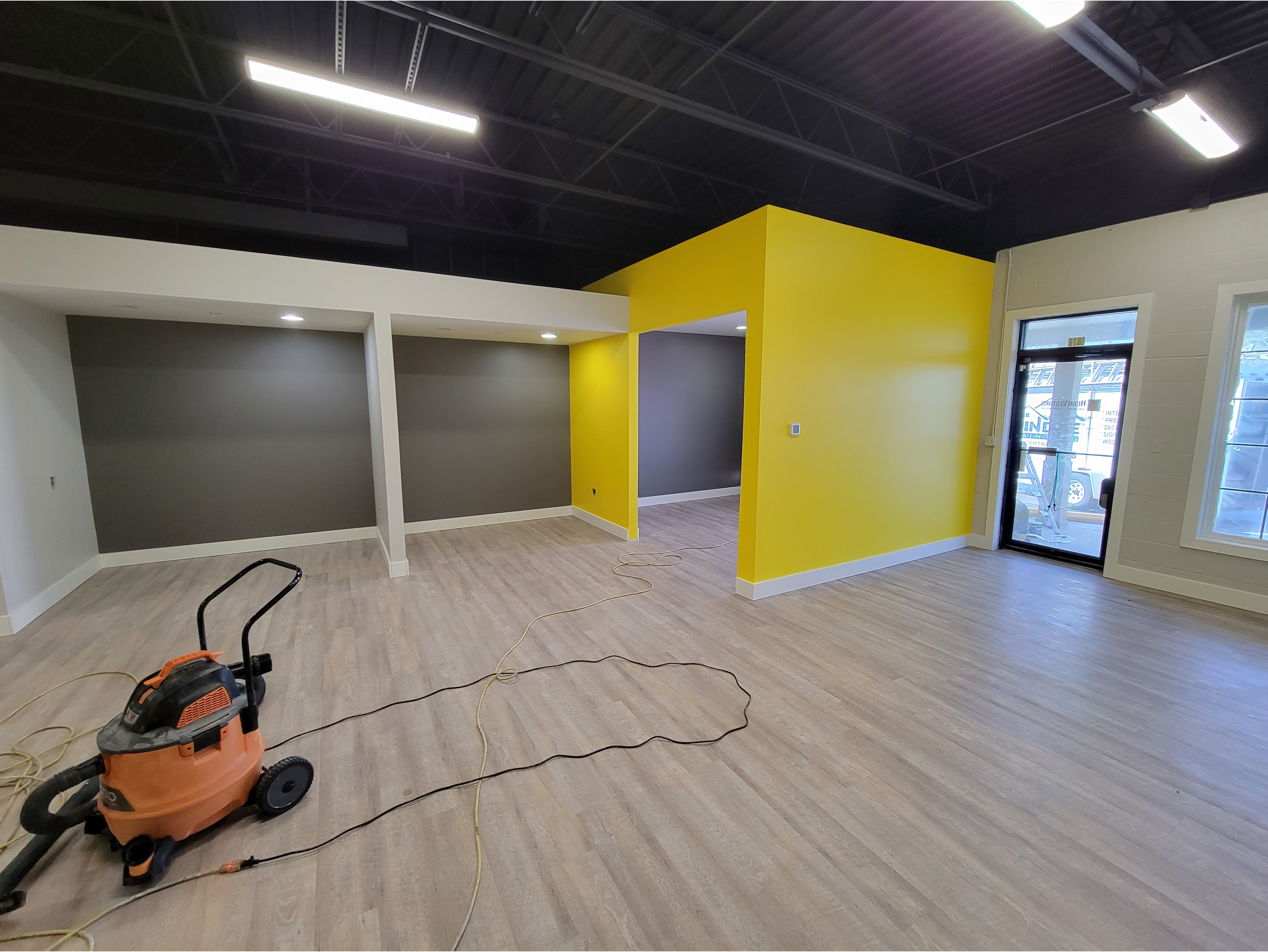 A commercial interior space featuring a modern paint job with a bold yellow accent wall and contrasting gray and white sections. The space has high ceilings, bright lighting, and new flooring, creating a clean and professional look. Perfect for showcasing commercial painting services and interior renovations for businesses.