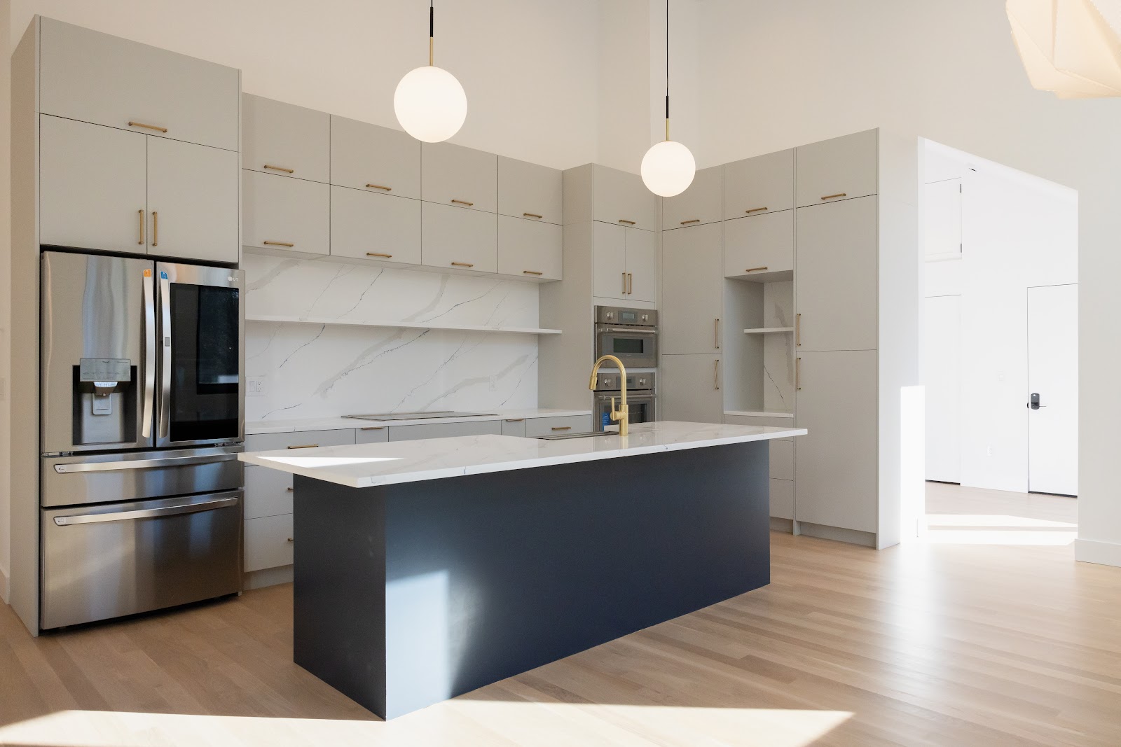 A modern, empty kitchen with a large island and stainless steel appliances. The kitchen features light gray cabinetry with gold hardware, a marble-patterned backsplash, and a dark blue island. Two pendant lights with frosted globe shades hang from the ceiling. The floor is made of light wood.