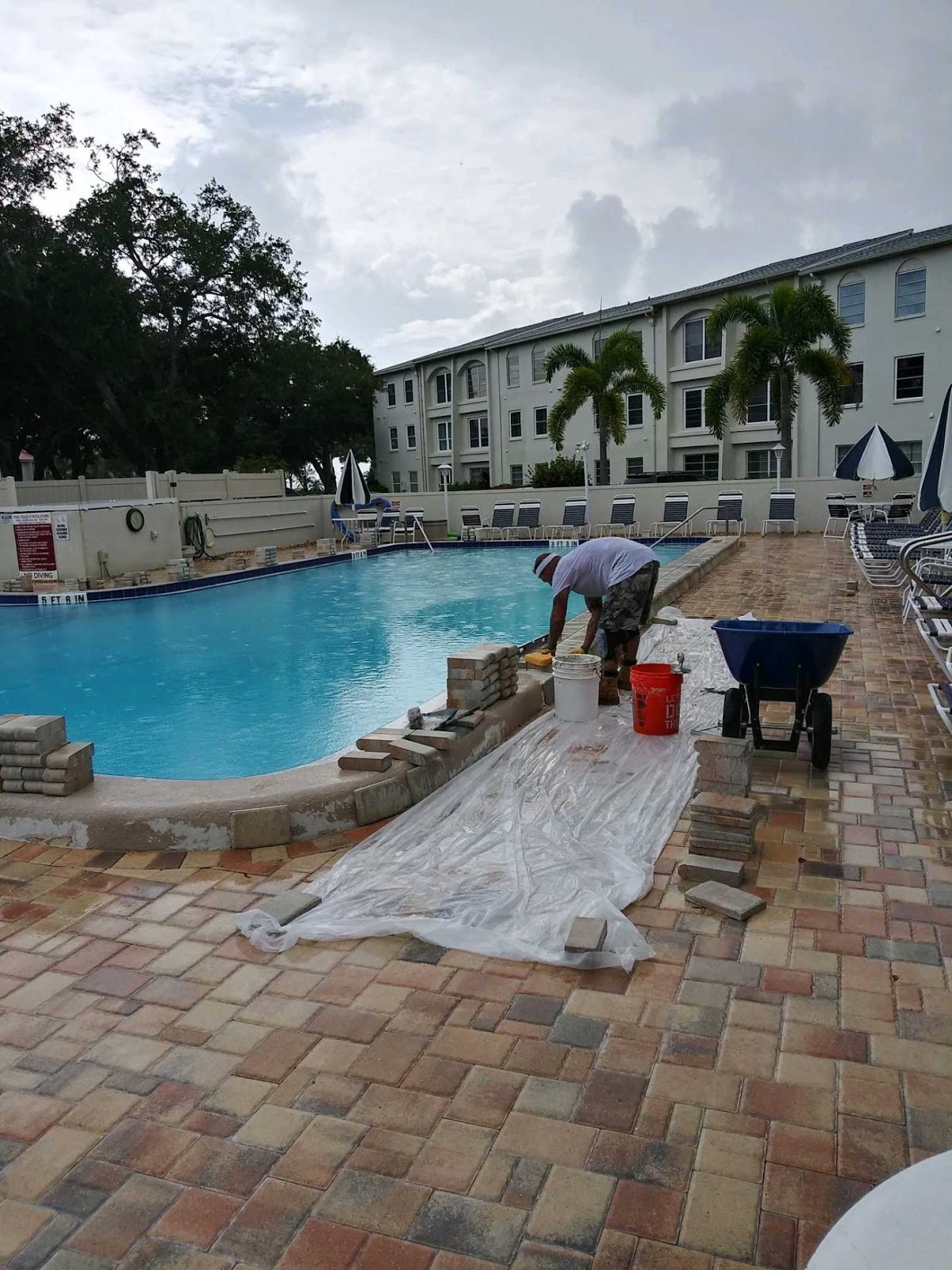 Commercial swimming pool renovation in progress with workers installing or repairing the surrounding paver deck.