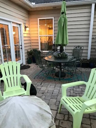 A cozy backyard patio with a round metal table, green chairs, a closed green umbrella, and paver flooring near a house.