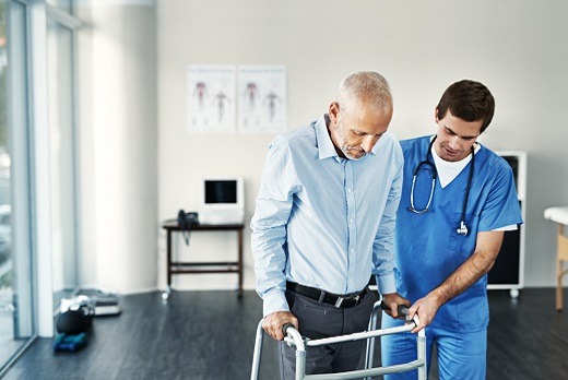 A professional caregiver walks beside an elderly woman using a walker in a bright hallway, offering guidance and encouragement. This image represents mobility support, fall prevention, and dedicated one-on-one care provided by 7 Sisters Homecare.