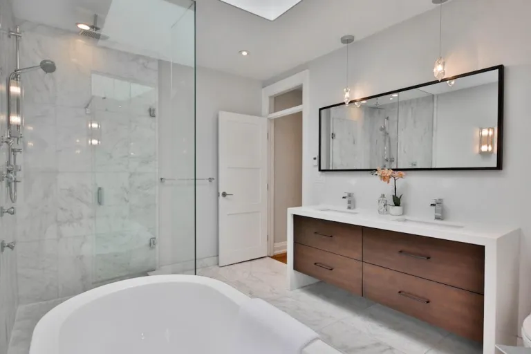 A modern bathroom with a freestanding bathtub, a glass-enclosed shower with marble walls, and a double vanity with a large mirror and modern pendant lights.