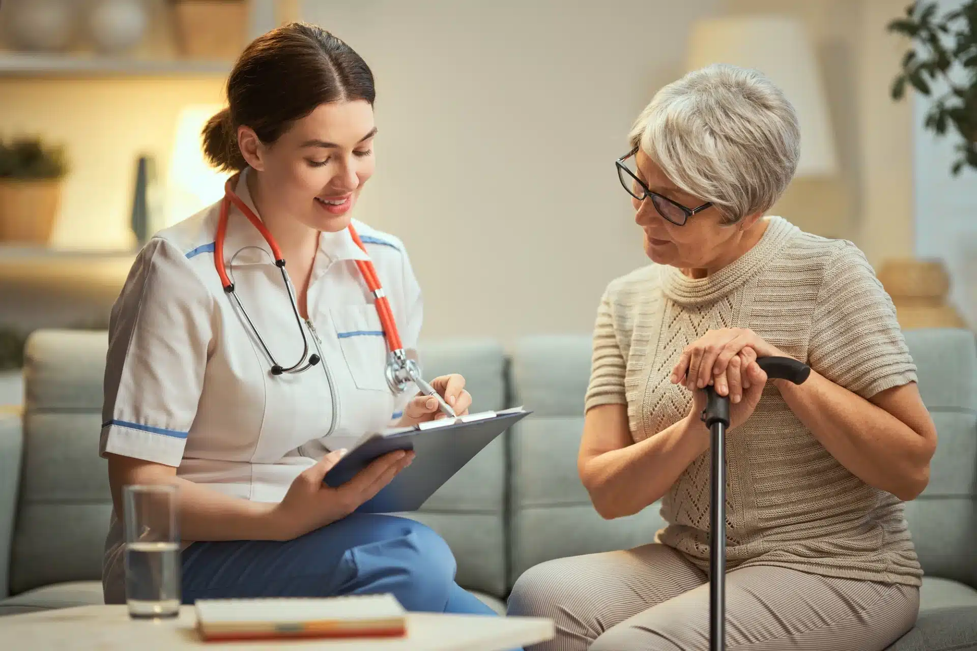 A smiling female caregiver in a white uniform and blue pants, wearing a stethoscope, is sitting on a sofa and taking notes on a clipboard. She is looking down at the clipboard with a friendly expression. Next to her, an elderly woman with gray hair, wearing glasses and a beige knitted sweater, is sitting on the sofa and holding a walking cane. The scene appears to be a home healthcare or medical consultation setting.
