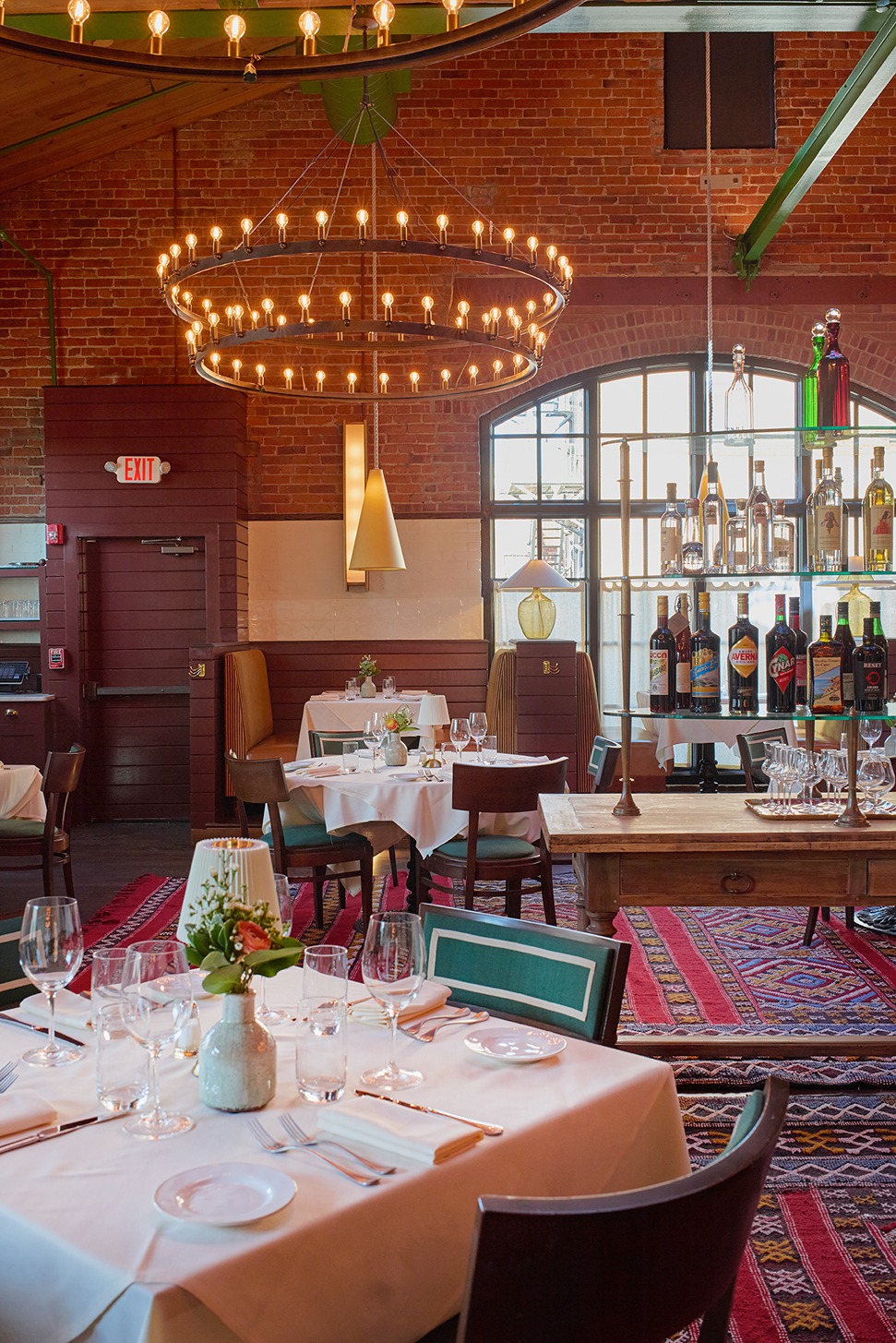 A spacious restaurant interior featuring exposed brick, red patterned rugs, and two large circular tiered chandeliers. Tables are set with white cloths, and a central wooden table holds a glass shelf of liquor.