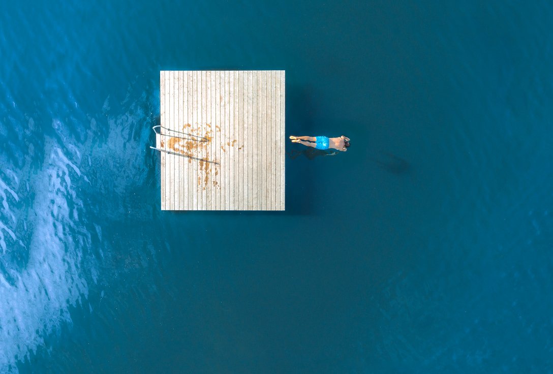 Overhead view of a person in blue shorts floating calmly next to a wooden dock in deep blue water, evoking a sense of tranquility and solitude.