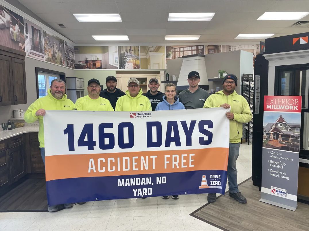 Eight team members stand inside a building materials showroom holding a large Builders FirstSource banner that reads “1460 Days Accident Free – Mandan, ND Yard.” Kitchen cabinetry, display fixtures, and product signage are visible in the background, including an “Exterior Millwork” display. The group is gathered to celebrate more than four years without a workplace accident.